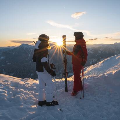 Two skiers in winter gear stand with skis on a snow-covered summit at sunrise in the mountains