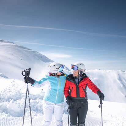 Two best ager female skiers hug and laugh on the slope, surrounded by snowy mountains, holding ski poles.
