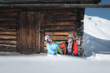 Two best ager female skiers laugh in front of a rustic wooden hut, enjoying the snow, sun, and alpine view.