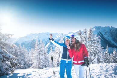 Zwei Skifahrerinnen machen ein Selfie am Pistenrand vor verschneitem Alpenpanorama, lachen und genießen die Sonne.