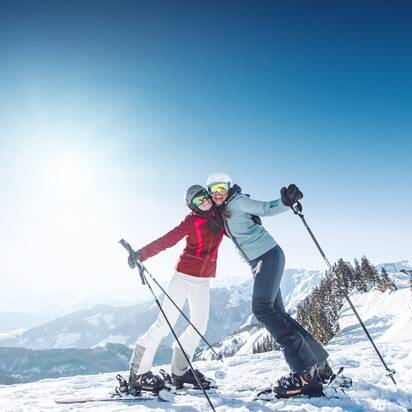 Zwei Skifahrerinnen umarmen sich auf der Piste, tragen Skibrillen und Helme, genießen das Alpenpanorama im Hintergrund.