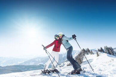 Zwei Skifahrerinnen umarmen sich auf der Piste, tragen Skibrillen und Helme, genießen das Alpenpanorama im Hintergrund.