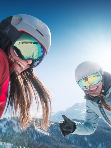 Two female skiers laugh wearing helmets and ski goggles, enjoying the sun and snowy Alps, with ski slopes behind.