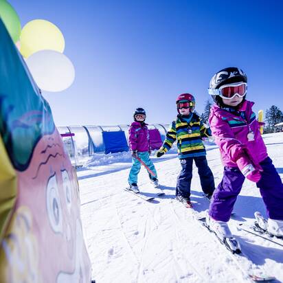 Drei Kinder in bunten Skianzügen fahren auf einer sonnigen Skipiste zwischen Skischule-Aufbauten, lachend und motiviert.