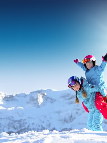 Two girls in blue ski outfits play and laugh in the snow with snowy mountains in the background, one raising hands.